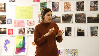 a teacher speaks at front of room with an artwork-covered wall in the background