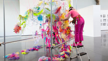 a student installs a piece suspending from the ceiling that is made up of colorful scraps of fabric