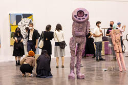 a tall, robotic humanoid sculpture stands in the center of a gallery surrounded by people and other artworks