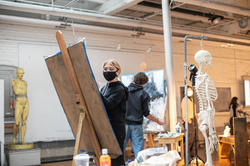 a student, facing the camera, draws on a large piece of newsprint next to a skeletal model and another drawing student