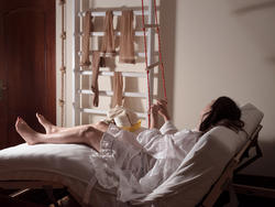 a person in a white dress sits on a reclined futon while holding a rope ladder inside a white room with wooden door and pantyhose hanging from a clothing rack