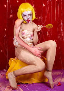 a nude figure with a yellow wig and painted mask on their face sits on a yellow chair in front of a red background