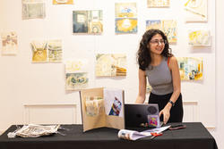 Reem Al Ani stands and smiles at a table of her work with paintings hung up on the wall behind her