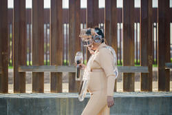 Women in scuba like suit and glass head piece walking in front of the Mexican-American border wall.