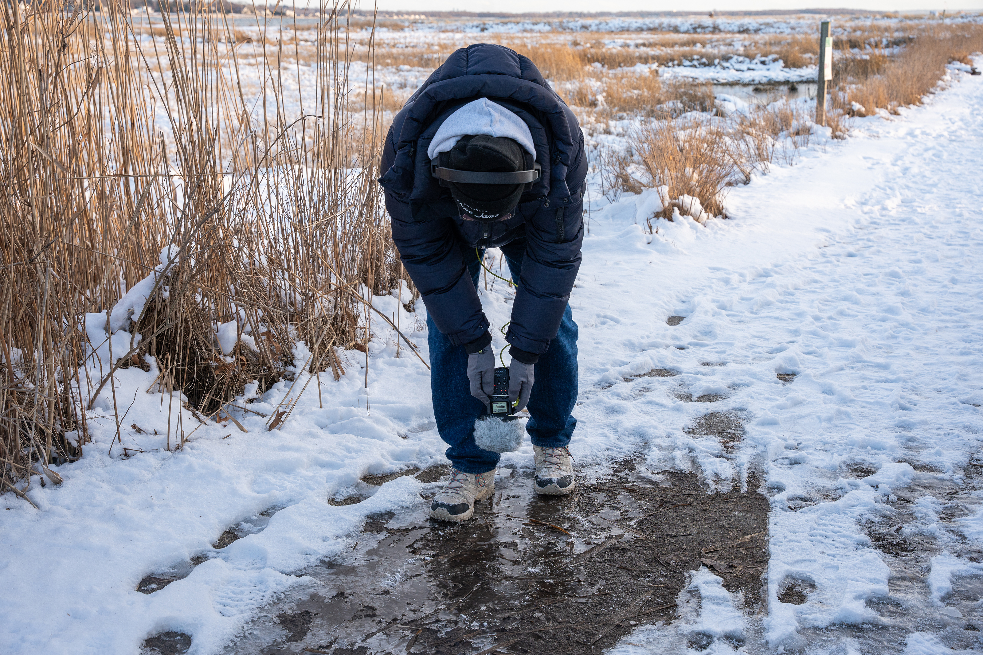 a student bends over to record the sound of their shoes cracking ice at the beach