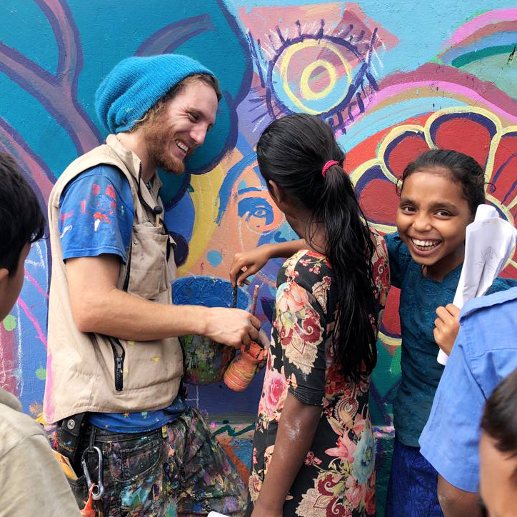 Dreadlocked and colorfully dressed Max Frieder helps kids in a refugee camp paint a mural.