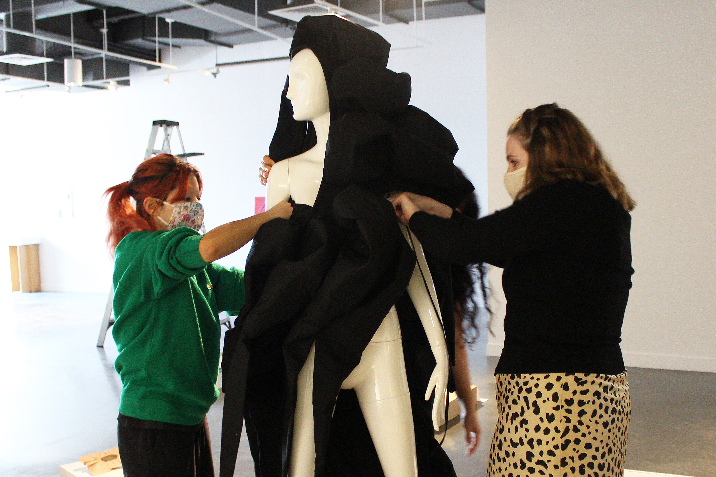 Two young women wearing masks drape a garment on a mannequin.