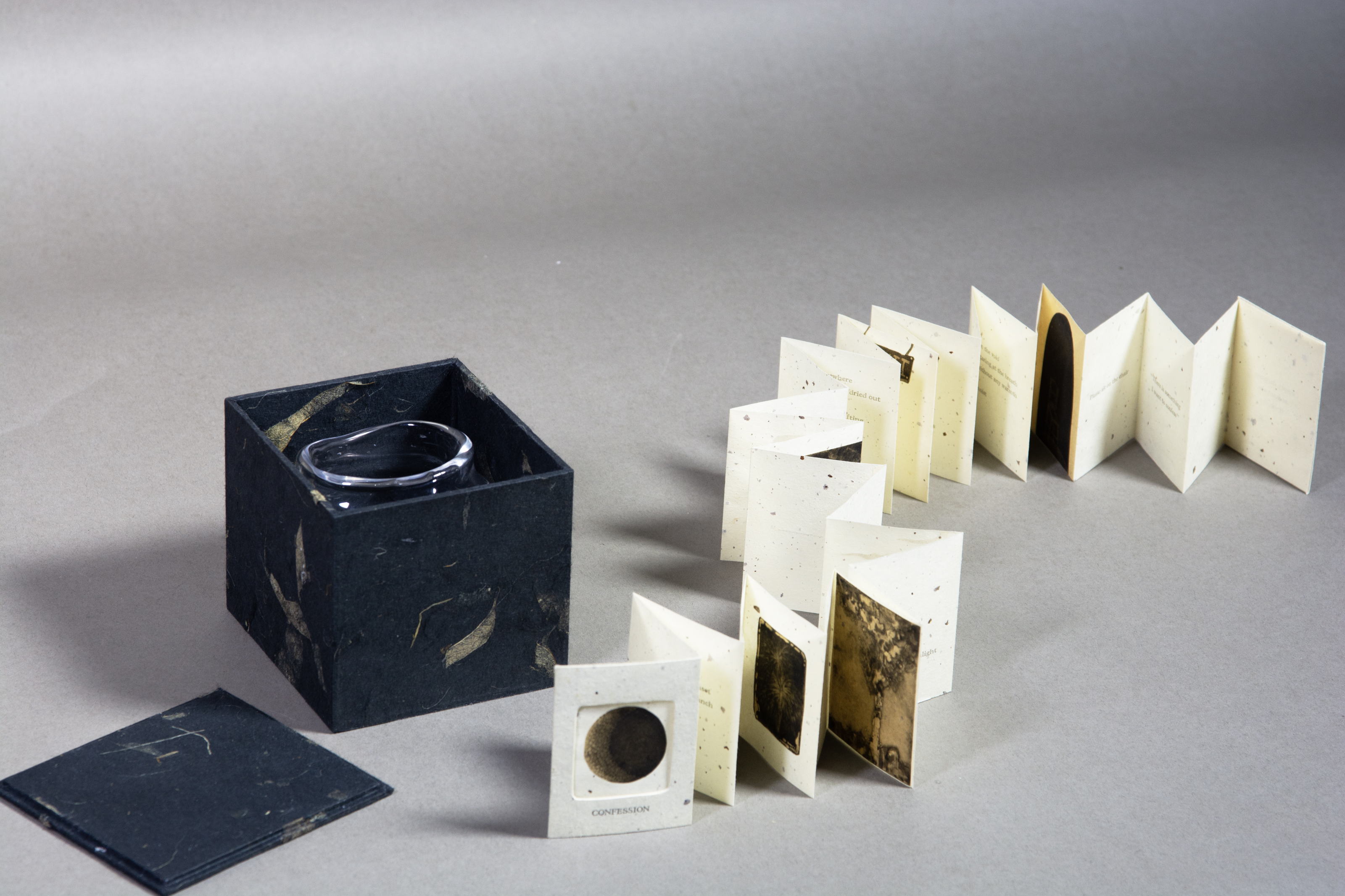 A small accordian booklet spread out next to a blue box with a handmade glass jar inside designed by Shihan Zhu