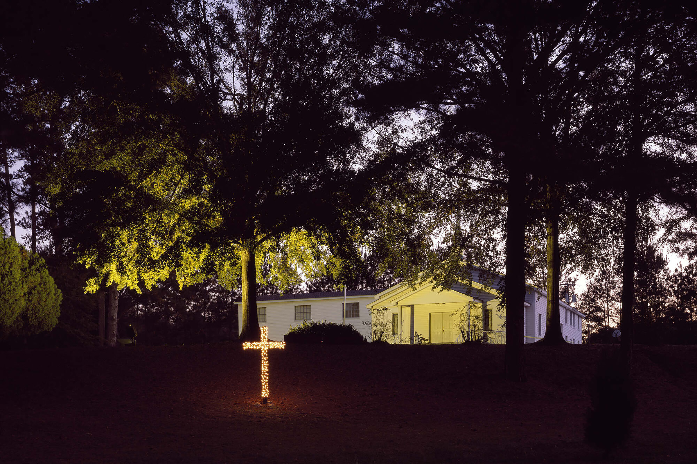 image of a house at dusk with a cross covered in small white lights burning on the lawn