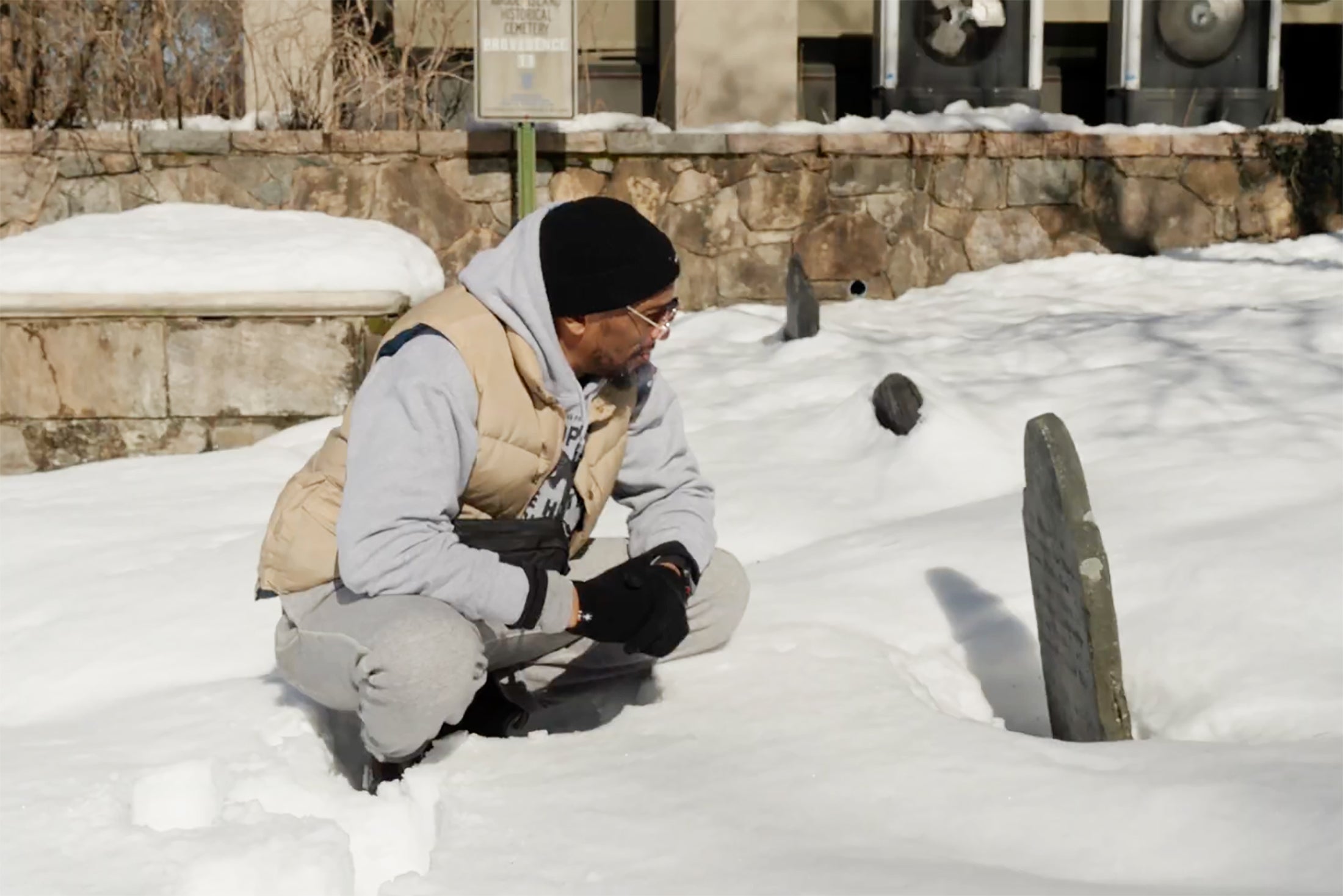 educator Marco Williams squats down to read a gravestone in a snow-covered cemetery