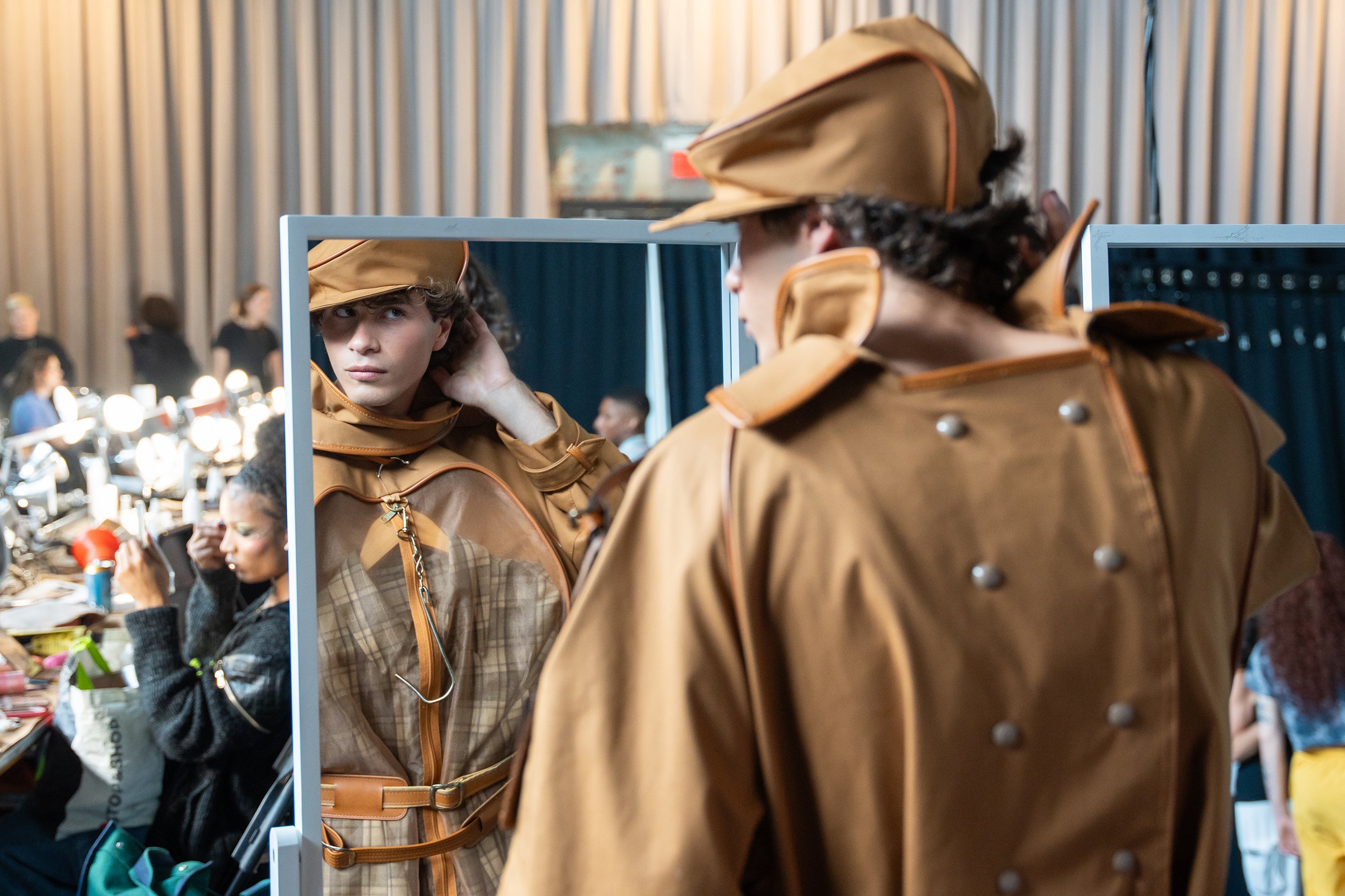 a model wearing a suit fashioned out of a hanging suitcase checks his hair in the mirror backstage