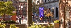 a blue flag with the letters RISD hangs in downtown Providence as students walk near green and gold foliage
