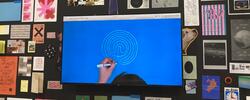 a monitor with a hand drawing a white spiral on a blue surface, surrounded by multicolored prints