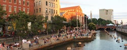 a large number of people sit on steps along a stretch of river running through downtown Providence