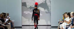 a model in a black, 19th-century military-style uniform (with red belt and hat feather) walks a runway with audience sitting on left and right