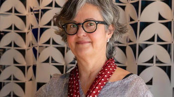 Image of a woman with short grey hair and glasses wearing a bright red beaded necklace and a grey shirt in front of a tapestry with cream colored arched shapes against a brown background