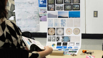 a student's hand reaches towards a long sheet with many small, square images hangs from a wall, surrounded by other printed works