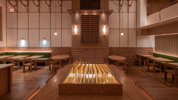 a golden sculptural structure atop a table at the center of a lightly-lit room with many wood tables atop a dark hardwood floor