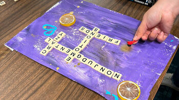 A person is working on an artwork with citrus slices and crossword pieces on a purple background.
