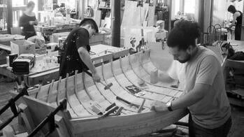 two students build a boat-like structure inside a design studio with other students working in the background