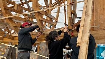 three students work outdoors on constructing a wooden boat