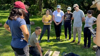 students stand on a grassy field around an instructor crouched over a saw and pieces of cut wood