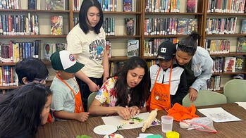 five students surround a teacher at a library table working with several pieces of colored paper, with full library shelves behind them