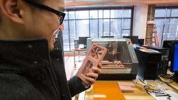 a student holds a smartphone-sized object nearby a 3D printer
