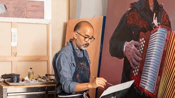Vincent Valdez holds a paintbrush while standing alongside a large-scale painting of a man holding an accordion