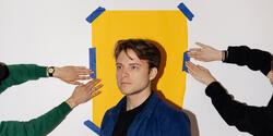 Bentel stands in front of a yellow piece of paper as hands tape it to the wall