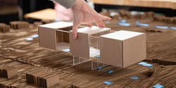 a hand lays a cardboard model of a building onto a cardboard model of a street grid