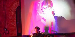 Patrick Buhr in pink lighting with face on screen behind him