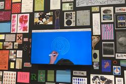 a monitor with a hand drawing a white spiral on a blue surface, surrounded by multicolored prints