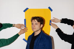 Bentel stands in front of a yellow piece of paper as hands tape it to the wall