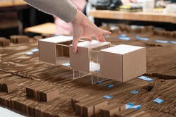 a hand lays a cardboard model of a building onto a cardboard model of a street grid