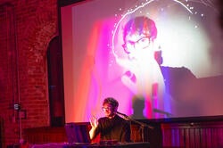 Patrick Buhr in pink lighting with face on screen behind him