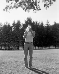 black and white photo of a person holding a jar as they walk along grass with trees in the background and gnats hovering around the person