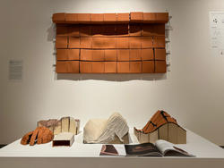 several clay sculpted clay structures rest on an exhibition display stand, with another reddish-brown structure, like a small roofed exterior, hanging overhead