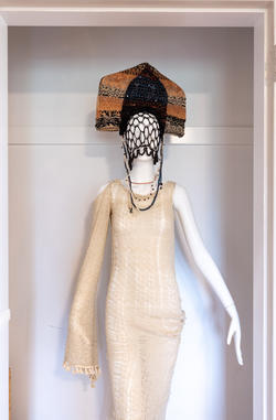 a mannequin outfitted with a cream-colored knit dress, multicolored headpiece and netted face covering