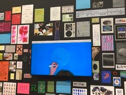 a monitor with a hand drawing a white spiral on a blue surface, surrounded by multicolored prints