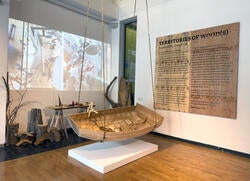 a wooden boat structure suspended in air inside a gallery space, with a video projection in the background