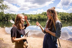 a student holds a small flora specimen for another student to view while standing on a beach with water, trees, and clouded blue sky in the far background