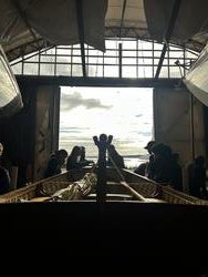 students stand around boat inside darkened boat shelter, with light coming in through doorway behind them