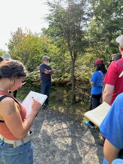 students stand outdoors around a person, taking notes as they speak