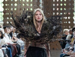 a model wears an outfit with a bird nest-inspired object with a runway audience in the background