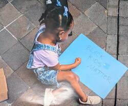 A young Black girl with two blue bows in her hair wearing denim overalls and a lavender shirt sits on a paved stone surface and draws on a blue poster board that reads "Black Kids Matter" in all caps.