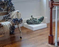 a sculpture of a fantastical green creature rests on a short gallery podium, flanked by other artworks inside a gallery