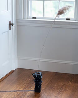 an irregularly shaped black cylinder, sprouting a wire topped with a feather, stands on a wood floor near a well-lit window