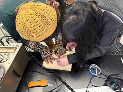two students lean over a small device while flanked by sound equipment