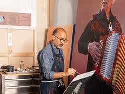 Vincent Valdez holds a paintbrush while standing alongside a large-scale painting of a man holding an accordion